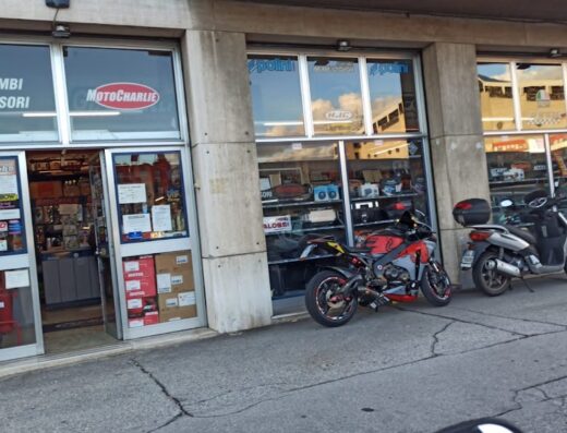 Negozio Moto Charlie con vetrine che espongono ricambi e accessori per moto e scooter, con moto parcheggiate davanti.