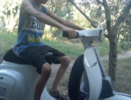 Ragazzo sorridente su una Vespa d'epoca bianca in Sicilia, un'esperienza di viaggio unica su due ruote con il Moto Club d'Epoca Sicilia.
