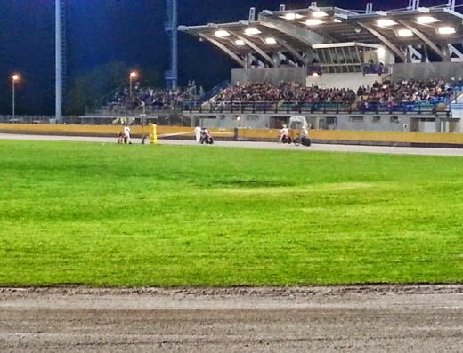 Vista del Moto Club Olimpia con pista verde, tribune affollate e moto sul circuito durante un evento serale.