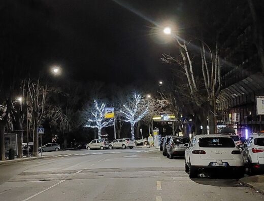 Strada notturna illuminata con alberi decorati, auto parcheggiate e semafori, atmosfera urbana serale a Genova, ideale per moto touring.
