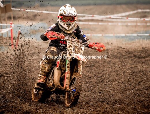 Pilota di motocross in azione durante un evento di Moto Day, su una pista fettucciata e fangosa. Casco, occhiali e protezioni completano l'equipaggiamento per questa disciplina di enduro.