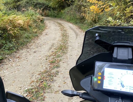 Moto enduro in Abruzzo: vista dal manubrio di una moto da viaggio su strada sterrata immersa nel verde autunnale. Accessori e display visibili.