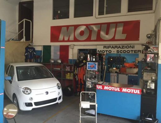 Interno di un'officina moto con una Fiat 500 bianca parcheggiata, insegna Motul, attrezzature per riparazioni moto e scooter.