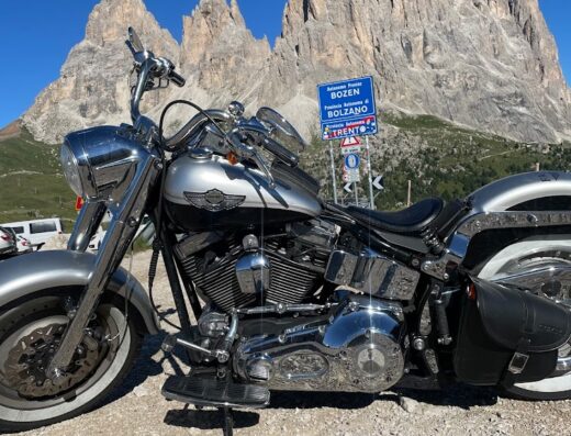 Moto Mozzini: una motocicletta Harley-Davidson argento parcheggiata con le Dolomiti e cartelli stradali di Bolzano e Trento sullo sfondo.