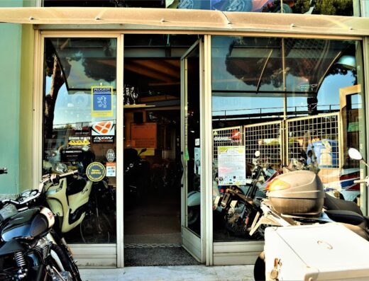 Ingresso del negozio Moto Nois S.A.S. con diverse moto parcheggiate e vetrine ricche di adesivi di marchi motociclistici.