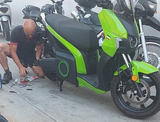 Moto Officina Due Ruote: Un uomo lavora su uno scooter elettrico verde brillante in un'officina, con altri scooter e moto parcheggiati sullo sfondo. Servizio di riparazione moto.