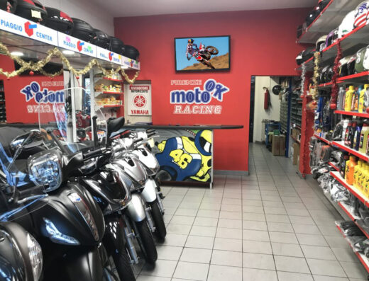 Interno del negozio Moto Ok Firenze con esposizione di scooter, caschi, ricambi e accessori moto. In evidenza il logo del negozio e un televisore con immagini di motocross.