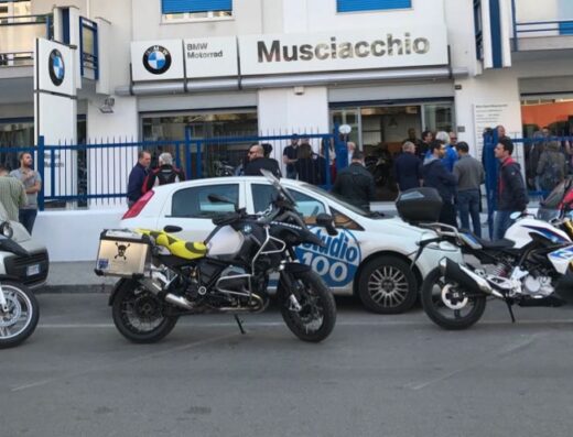 Foto di Moto Sport Musciacchio Srl, concessionaria BMW Motorrad, con diverse moto parcheggiate di fronte e un gruppo di persone riunite all'esterno.
