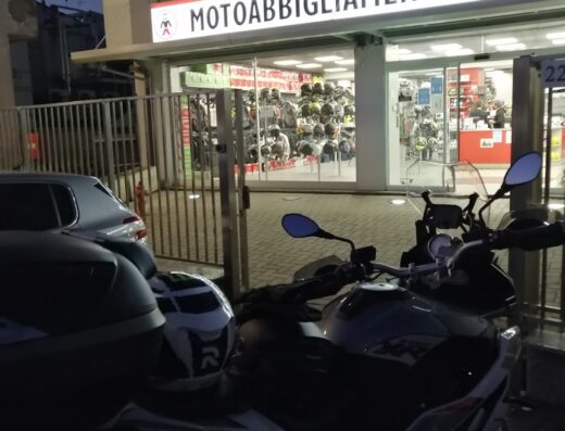Moto parcheggiata di fronte al negozio Motoabbigliamento.it con vetrina piena di caschi e accessori per motociclisti.