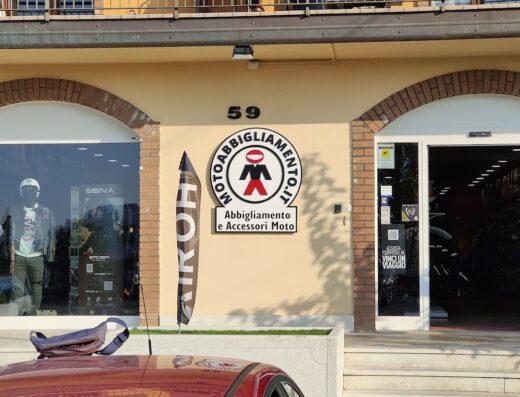 Facciata del negozio Motoabbigliamento con insegna e vetrine che espongono abbigliamento e accessori moto, focus su Airoh e Sena. Auto rossa parcheggiata.