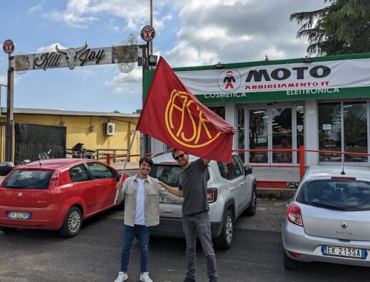 Due persone davanti a un negozio di motoabbigliamento con una bandiera, auto parcheggiate, insegna Kill Joy, cielo nuvoloso.