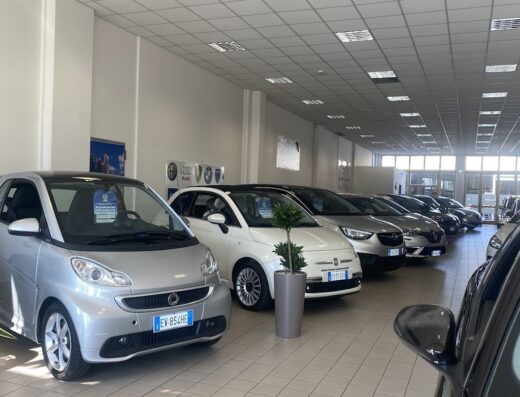 Interno di concessionaria auto con esposizione di diverse vetture, tra cui Smart, Fiat e Opel, in vendita. Servizi di moto e auto sport.