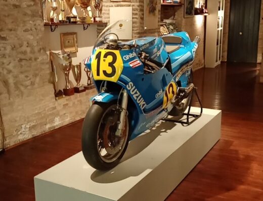 Motocicletta Suzuki da corsa numero 13 esposta su un piedistallo bianco in un museo, trofei sullo sfondo. Moto Club Franco Uncini.