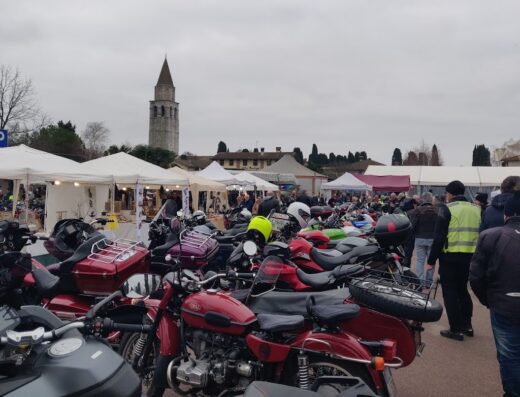 Affollato raduno di motociclette del Motoclub Morena, con diverse moto parcheggiate e stand visibili, sullo sfondo un campanile.