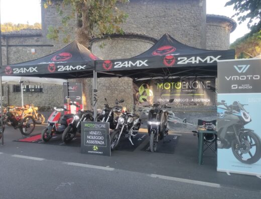 Esposizione di moto e scooter elettrici MotoEngine durante un evento all'aperto, con tende 24MX, cartelli pubblicitari VMoto Super Soco, vendita, noleggio e assistenza di veicoli elettrici.
