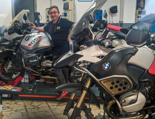 Motofficina Ingallina: un meccanico sorridente posa accanto a due moto BMW GS in officina a Catania, con attrezzature e ricambi visibili.