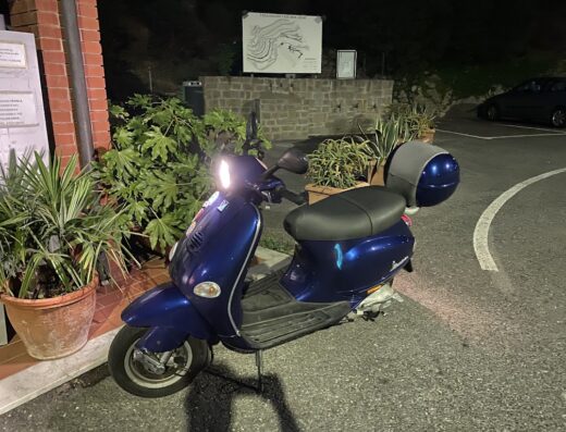 Scooter blu parcheggiato di fronte a un edificio, ideale per esplorare le strade con stile. Accessori moto e soluzioni di viaggio disponibili su Motofollie di Enzo Barone.