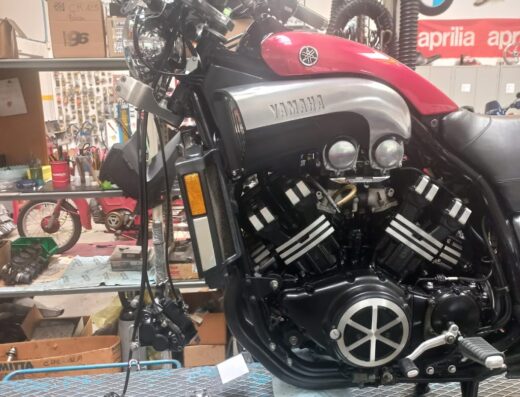 Motocicletta Yamaha V-Max rossa e argento su un banco da lavoro in un'officina, con accessori e attrezzi visibili sullo sfondo.