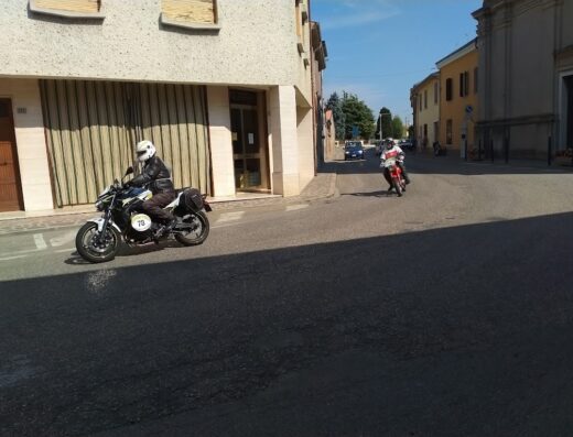 Motogiro d'Italia: Un motociclista su una moto moderna accessoriata con borse laterali percorre una strada cittadina durante il tour, mentre un altro motociclista su una moto rossa lo segue a distanza.