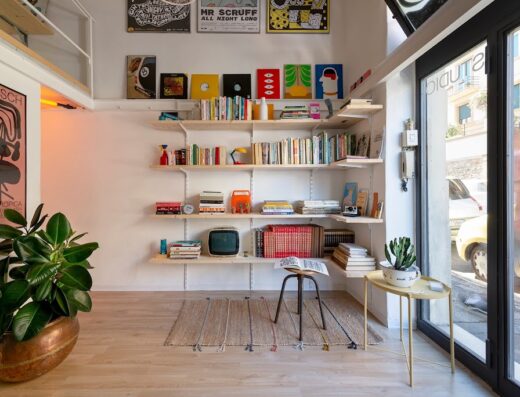 Interno di uno studio con libreria a muro piena di libri, dischi e oggetti decorativi, una pianta in vaso in primo piano e vista sulla strada attraverso una vetrata.