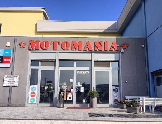 Foto del negozio Motomania, rivenditore di moto e accessori, con insegna ben visibile. Presenti anche marchi come Vespa, Aprilia e Voge. Ampia vetrina e indicazioni per officina e accettazione.