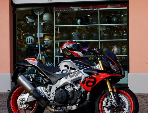 Una moto Aprilia parcheggiata di fronte al negozio Motone Racing con un casco appoggiato sopra. Accessori e abbigliamento per motociclisti in vendita.