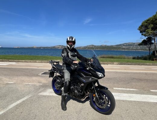 Un motociclista in sella a una Yamaha Tracer nera con dettagli blu, indossa casco e abbigliamento protettivo, sullo sfondo mare e costa della Sardegna.