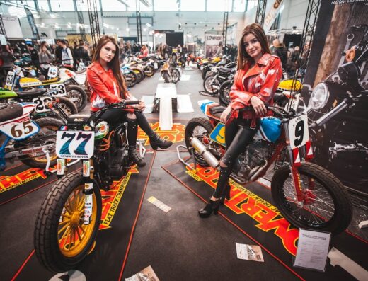 Due ragazze in giacca di pelle rossa posano su moto customizzate al Motor Bike Expo, con altre moto sullo sfondo.