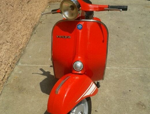 Una Vespa rossa d'epoca parcheggiata su un marciapiede, con altre moto e scooter sullo sfondo. Ideale per il marketplace di moto e accessori.