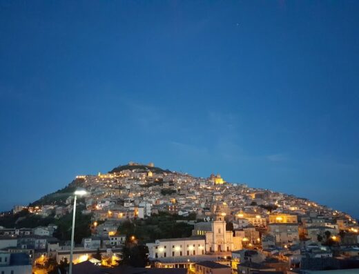 Veduta notturna di Agira, Sicilia, con case illuminate sulla collina e cielo blu scuro, ideale per viaggi e moto tour.