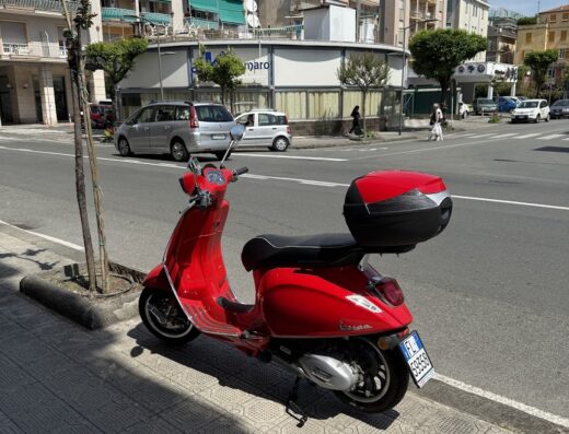 Vespa rossa parcheggiata in strada con bauletto portapacchi, ideale per il noleggio scooter e moto in città.