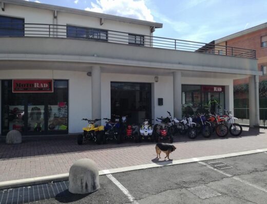 Esposizione di moto e quad presso il negozio Motorad1, con un cane in primo piano. Vetrina di accessori moto visibile.