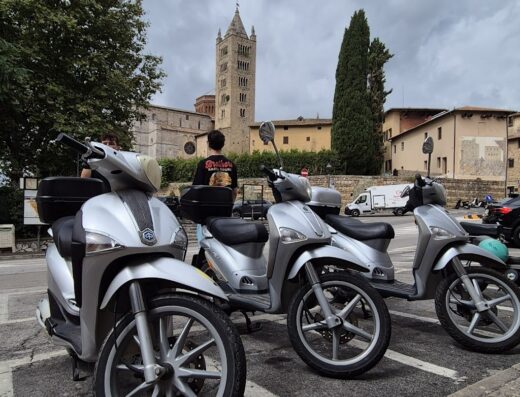 Tre scooter Piaggio Liberty parcheggiati a San Miniato, Toscana, con la torre campanaria romanica sullo sfondo, tipico paesaggio italiano.