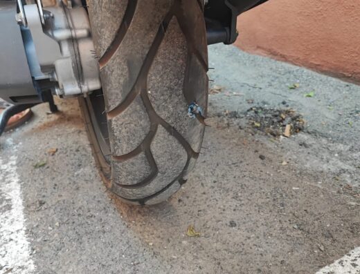 Dettaglio della ruota posteriore di una moto di Incerti Massimiliano, che mostra uno pneumatico usurato con un potenziale danno, parte del motore e dello scarico visibili.