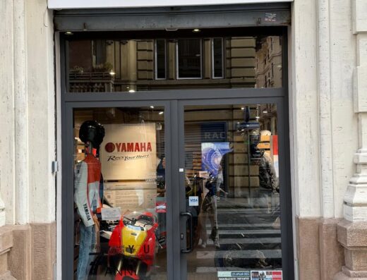Motorcity Genova: vetrina del negozio con moto d'epoca rossa e gialla esposta, abbigliamento Yamaha e cartelli informativi. Accessori e marketplace per motociclisti a Genova.