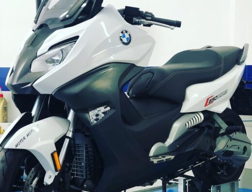 Una moto BMW C 650 Sport bianca e nera su una piattaforma blu in un'officina, ideale per viaggi e accessori moto.