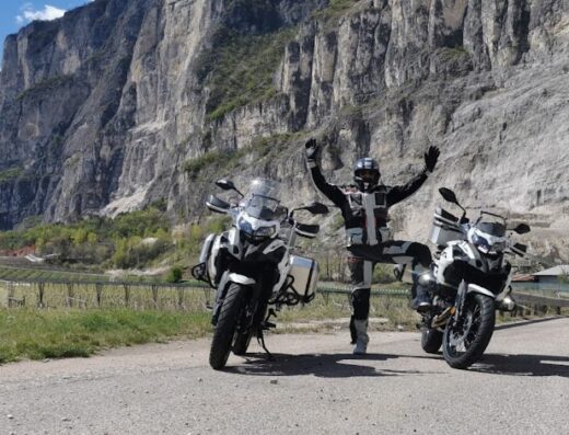 Noleggio moto in Alto Adige: motociclista felice tra due moto BMW GS davanti alle montagne e ai vigneti del Sudtirolo.