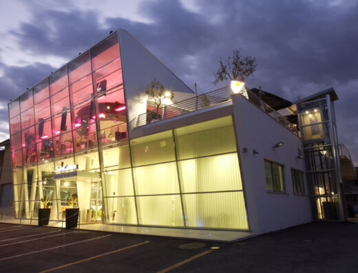 Edificio moderno di Motorsport SRL con facciata in vetro illuminata, design architettonico innovativo, illuminazione interna rosa e gialla, spazio espositivo per moto e accessori, servizi per motociclisti.