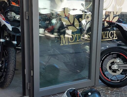 Motoservice GmbH: caschi moto bianchi e neri davanti all'ingresso con vetrine che riflettono le moto all'interno, officina specializzata e accessori per motociclisti.