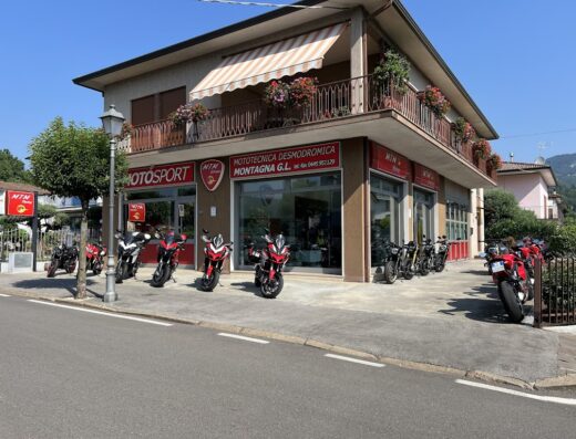 Negozio Motosport Montagna MTM Racing con diverse moto esposte all'esterno, vetrine con insegne, balcone fiorito e cartelli pubblicitari del marchio.