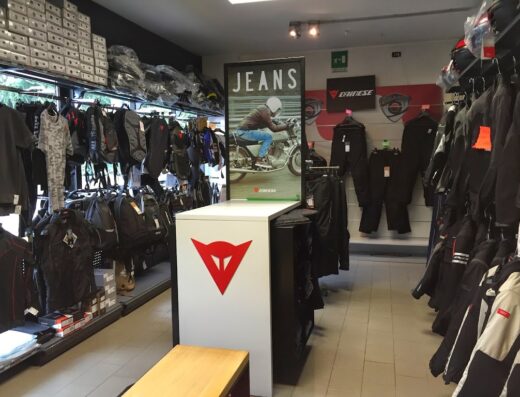 Interno di un negozio di abbigliamento e accessori moto: giacche, pantaloni, caschi e zaini esposti, con cartello "Jeans" e logo Dainese.