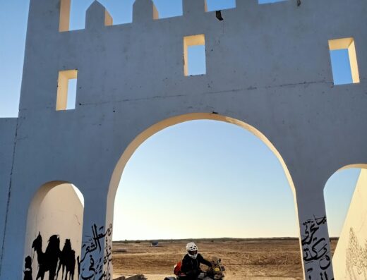 Motociclista in viaggio avventura in Marocco, attraversa un arco decorato con graffiti di cammelli e scritte arabe. Tour operator Motovacanze offre viaggi e servizi per motociclisti.