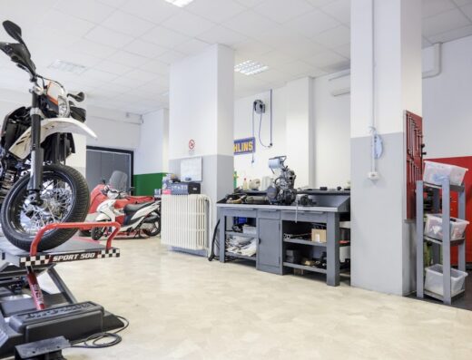 Moto da cross bianca su piattaforma elevatrice in officina con banco da lavoro e altre moto sullo sfondo. Servizi di riparazione moto e accessori.