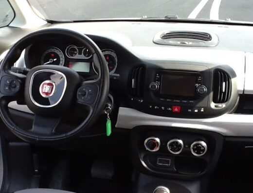 Interni di Fiat 500L bianca con volante, cruscotto, cambio manuale e sedili bicolore, ideale per noleggio auto e viaggi su strada.