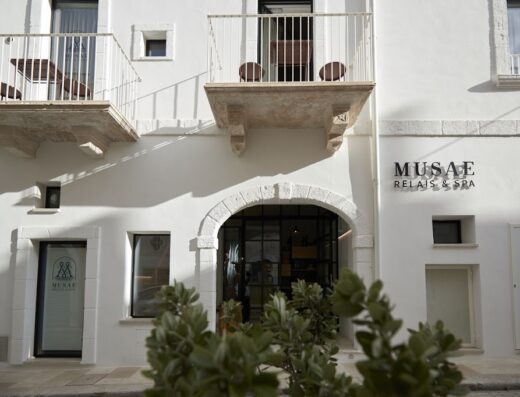 Facciata bianca del Musae Relais & SPA con balconi in pietra, ingresso ad arco e vegetazione in primo piano, che invita al relax e al benessere.