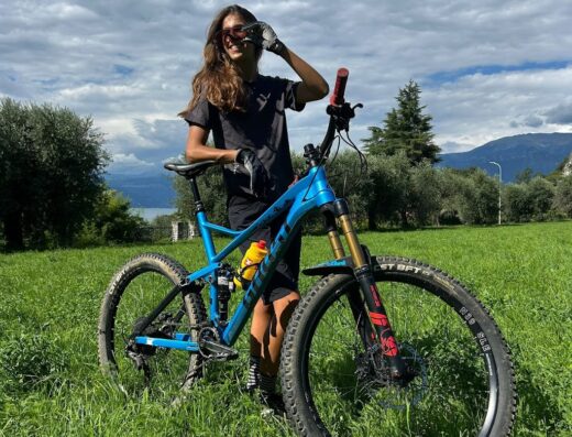 Una donna sorridente, con occhiali da sole, appoggiata a una mountain bike blu su un prato verde. Noleggio bici premium e tour all'aperto.