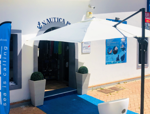 Ingresso del negozio Nautica Più, con servizi di noleggio barche e vendita accessori nautici. Insegna con logo e ancora, bandiera Seac Sub, ombrellone bianco e indicazioni per tour privati e taxi boat per le Isole Eolie.