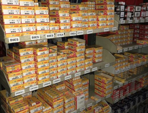 Scaffalatura piena di scatole di candele NGK di vari modelli (B6ES, B7ES, B8ES, B9ES, CR7HSA, ecc.) in un negozio di ricambi per moto. Accessori e componenti per motocicli.