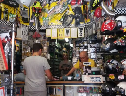 Interno del negozio Neroni Ricambi, con un cliente al banco, fornito di accessori e ricambi per moto, caschi e abbigliamento tecnico.
