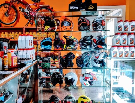 Interno negozio Nicolardi Moto con moto d'epoca rossa appesa al muro sopra una vetrina piena di caschi da moto di diversi colori e marche e scaffali con flaconi di olio motore e accessori per motociclisti.
